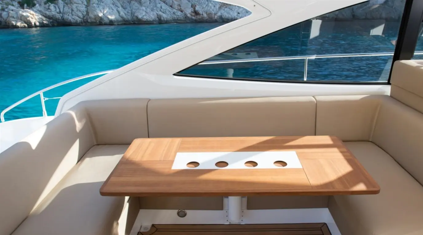 Stylish boat seating area with a wooden table featuring four cup holders, overlooking clear blue water and rocky shoreline.